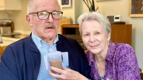 A head and shoulders photo of Ray and Anne Scowen. Anne, with short grey hair, is on the right of the picture smiling to camera and is holding up a beaker of tea for her husband Ray to drink. Ray is on the left of the picture. He has short grey hair and is wearing glasses. You can see their kitchen in the background.