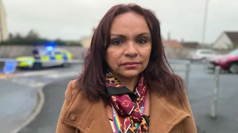 Ms Dossantos is wearing a brown coat and a patterned scarf. She's standing in the street. There is a police car behind her. She has brown eyes and shoulder-length hair.