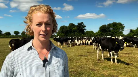 Dairy farmer stood in white checkered shirt. She has blonde hair, eye blues and is stood in front of cows standing in a field behind her.