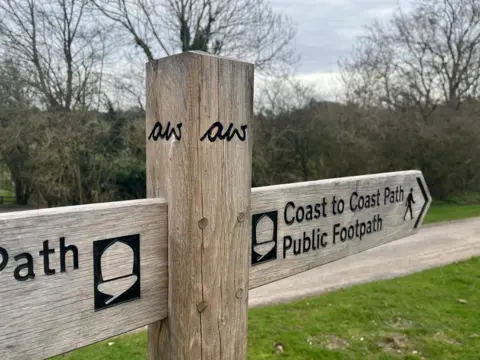 A wooden signpost stands at a junction of paths, with two horizontal arms pointing in different directions. The right‑hand arm reads “Coast to Coast Path, Public Footpath” with a walking‑figure symbol. The left‑hand arm shows part of the same acorn‑shaped National Trail marker. Trees and grassland fill the background, and a gravel path runs behind the sign. In the centre of the sign post are the initials aw carved into it.