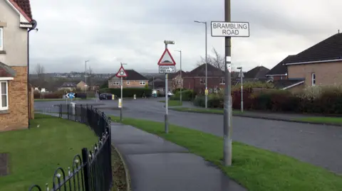 Road approaching a roundabout with modern houses on either side. There is a sign saying Brambling Road as well as one depicting speed humps and another a roundabout.