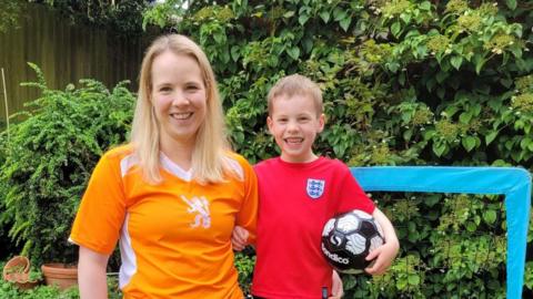 Emily Knight in a Holland football top and mid-length down blonde hair smiling next to her six-year-old son Edywn who is wearing an England top and holding a football in their garden