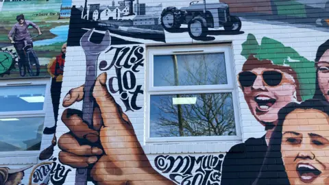 A segment of the mural that has a hand holding a wrench, a tractor, a person on a bike, black writing that says 'just do it', and three people smiling.