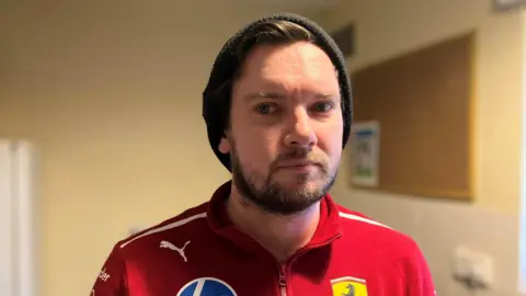 Shariqua Ahmed/BBC Andrew stares at the camera. He wears a red top with sports logos on it. A has a short beard and wears a black hat. The background is beige walls with a noticeboard to the right. 