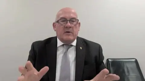 Richard Collier-Keywood appearing at the committee via video link. He is wearing a black suit jacket with a white shirt and grey tie and gesturing with both hands