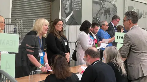 People standing in front of a table some holding clipboards and wearing rosettes. On the otherside with their backs to the camera are people sat down who will be counting and verifying votes.