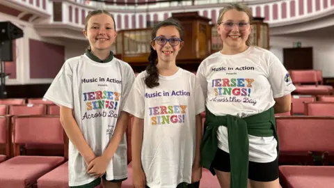 Three girls are smiling and wearing T-shirts saying Music in Action and Jersey Sings! They stand in front of chairs in St Helier Methodist Centre. 
