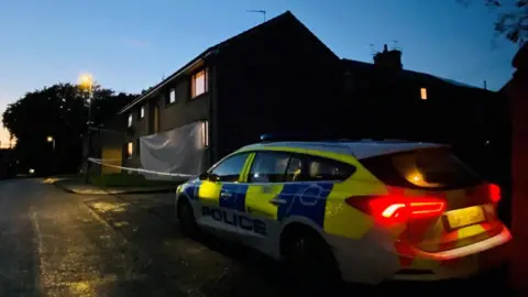 BBC Police car with lights on outside a house, and police tape.