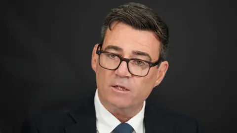 PA Media The prime minister, Andy Burnham, looks serious. The background is black, he is wearing a dark suit and black glasses