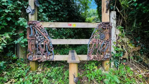 A stile through a hedge with recycled metal tools attached to each side