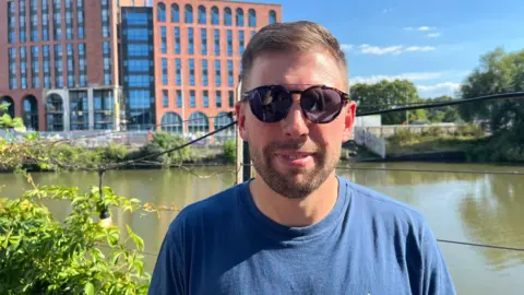 Dan Deeks, MD of Motion, in a blue t-shirt and wearing sunglasses, standing in front of a river, with buildings in the background