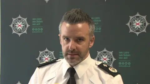 An officer is talking into mics. He's wearing a white shirt with black lapels.