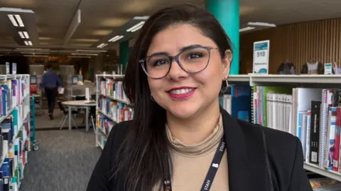 Gemma Dillon/BBC Image shows a lady with long brown hair wearing glasses.
She has a light brown roll neck jumper on with a black suit jacket. She is stood in front of rows of books in a library setting. 