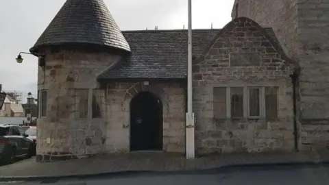 The outside of the former police station, which is a small stone-built structure with a turret-shaped section on the left and a flagpole in front of it.