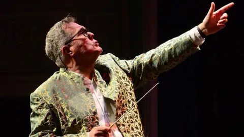 A conductor wearing a patterned green-and-gold jacket stands under stage lighting, holding a baton in one hand while pointing upward with the other. The background is dim, suggesting a concert hall or theatre setting.