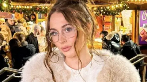 Amarni Jessica Rose Walton has her light brown hair tied back, with tresses hanging down. She is has glasses and is wearing a light-coloured fur, and white top, and is stood on stairs, in front of a busy Christmas party behind her.
