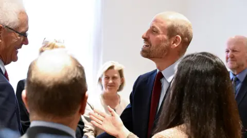 A group of people around one man who is smiling and wearing a suit. He is talking to those around him, who can mainly be seen from the back. 
