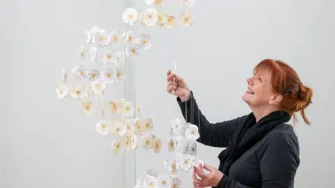 Jim Wileman Gillian dressed in black positions paper flowers that are hanging from the ceiling in an art gallery.