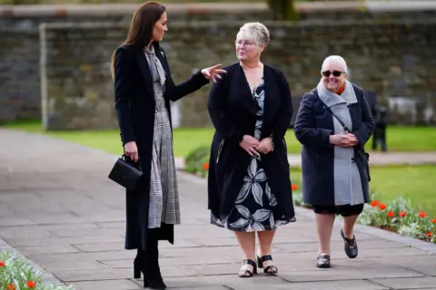 Getty Images The Princess of Wales speaking to two women in the memorial garden at Aberfan. A neatly-mown lawn and beds with red flowers can be seen in soft focus behind and to the right of the group. They are walking along a paving slab path, with a large stone wall in the background.