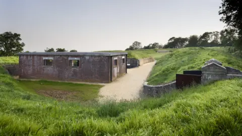 A red brick built low-level building sits in a grassy dip, a gravel path skirts around the front of the building. To the right of the pathway stand the main gates to the fort which is surrounded by trees.