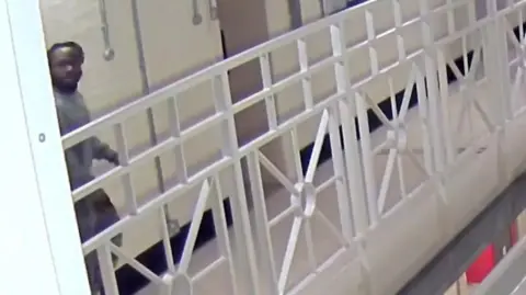 West Yorkshire Police/PA Media Screen grab taken from CCTV showing a prison corridor, with white metal railings and a staircase. A man in grey clothes is seen walking down the corridor and appears to be looking over his shoulder.