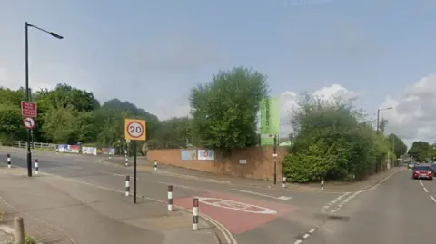 Street view of a junction in a suburban area. The road splits into two directions: one going uphill to the left with a 20 mph speed limit sign and bollards, and another continuing straight ahead marked as B6088. A brick wall with posters and a green sign is visible at the corner, surrounded by trees and greenery.
