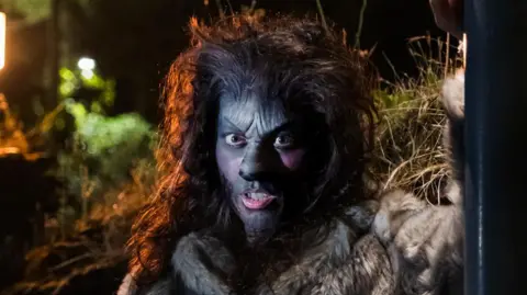 An actress in prosthetic makeup to make her look like a wolf. She wears a wig of long dark hair that has been teased and made to look big and bushy.