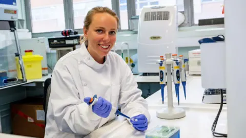 Dr Ellie Keeling seen wearing a white lab coat while sat in a lab. She is handling lab equipment with blue gloves.