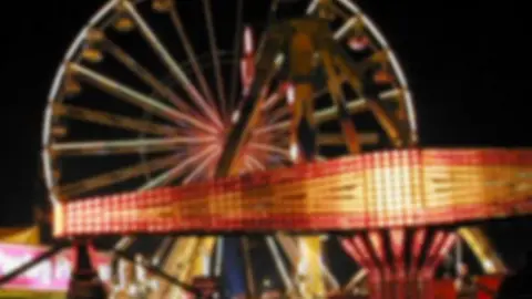 A blurred view of a funfair that has a Ferris wheel in the background. A diamond shaped red and yellow sign from a ride stands in front. 