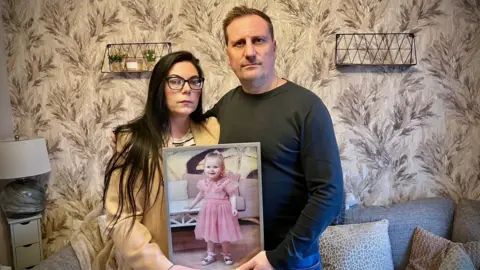 A man and a woman are standing in a living room holding up a picture of a young child with blonde hair. 