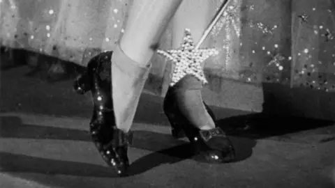 Metro-Goldwyn-Mayer/Getty Images A black and white close-up shot from the 1939 film showing Dorothy lifting up one heel while wearing the shoes, with a star-shaped wand pointing down at them. Glinda's full sequinned net skirt can be seen behind her