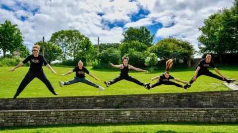 TUESDAY - Five young people wearing black T-shirts jump out from behind a stone wall in Swanage