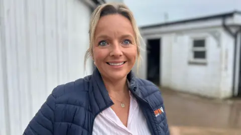 Katie Nairn with blonder hair tied back, wearing a pink striped shirt and wearing a blue padded jacket, smiling to camera and standing outside of a stable block 
