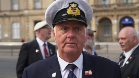 Thompson is wearing a dark navy suit jacket, a white shirt, and a dark tie.
A peaked white cap with gold-colored embroidered badge at the front.
Several small pins or badges are attached to the jacket lapels. One visible badge includes a small Union Flag design.