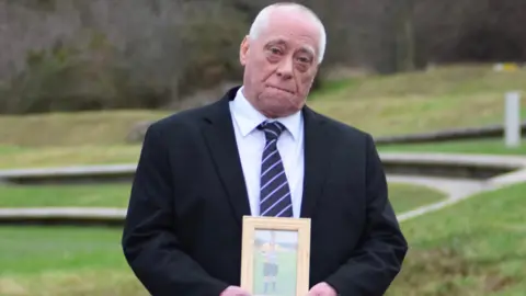 PA Media Alan Knox stands outside the Scottish Parliament wearing a suit with grass behind him holding a wooden photo frame with a picture of his son Lewis Knox inside. 