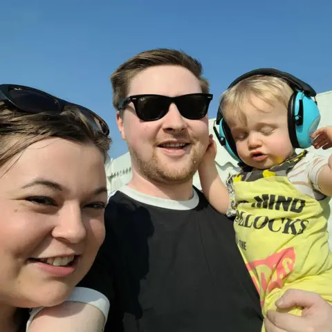 Family handout A man in a black t-shirt takes a selfie whilst holding a child wearing blue ear defenders, to the other side is a woman whose face is just above his arm