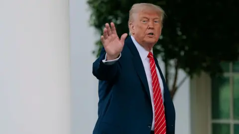 Donald Trump seen entering the White House and waving. He's wearing a suit with a red tie