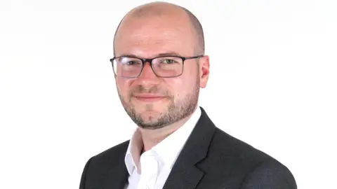 SaTH A corporate headshot of a man in a dark suit and open-necked shirt. He has a close-shaven head and short beard, and is wearing glasses.