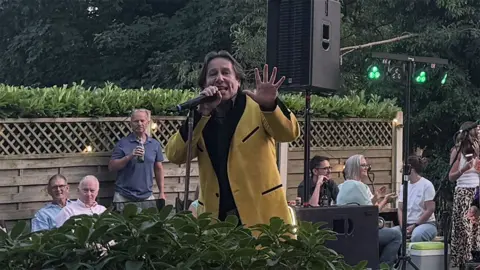 Showaddywaddy's singer in a yellow and black teddy boy suit jacket singing in the garden as people watch on