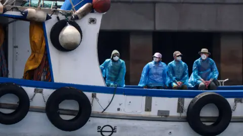 Reuters Four people are sitting on the back of a white boat with blue trim, and tyres hanging alongside. The four people are wearing blue protective raincoats and white facemasks.