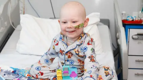Sarah Kent Vinnie, 4, is sitting on a hospital bed. He has no hair and has a tube going into his nose. Vinnie is smiling and wearing grey and blue pajamas with dogs on them. They are the dogs from a children's TV programme called Paw Patrol.
He has a few toys in front of him on the bed.