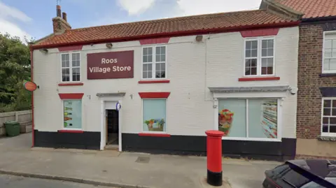 Google A village shop in a cottage-style brick building, the exterior painted cream and black. A red post box stands outside. Signs advertise "Roos Village Store" and "Post Office".