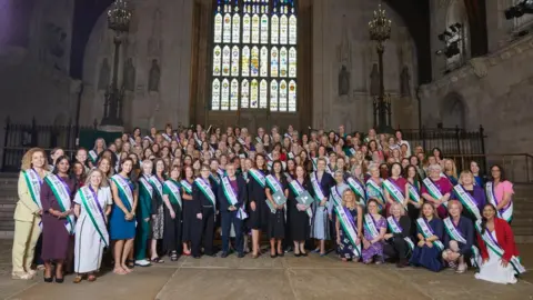 264 women MPs stand in Westminster Hall, all wearing the green, purple and white Centenary Action sashes. A stain glass tall window can be seen behind the group. 