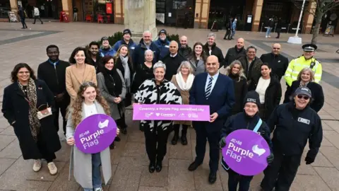 A group of around 30 people stand outside, with several holding purple signs saying 'Purple Flag' on them.