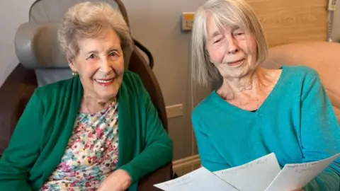 BBC Two older ladies sitting in chairs laughing, one wearing a multi-colour floral top and green cardigan, the other wearing a blue v-neck jumper