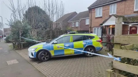 A police vehicle outside a property in Siskin Close. There are a number of properties and trees nearby, and police tape in front of the house. 