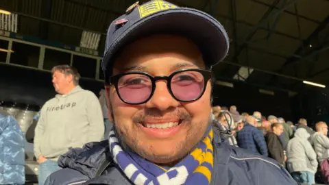 Stephen Huntley/BBC Nathan Doucette wears a navy baseball hat and jacket, with a scarf around his neck. He is sitting in the stand and grinning.