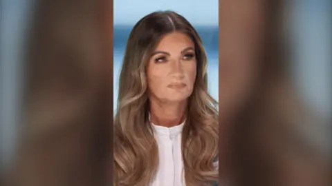 Karren Brady has long brown wavy hair and wears a white shirt. She looks stern.