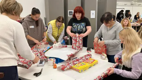 Volunteers standing round a table indoors wrapping gifts to be sent to Ukraine. They are all women, and on the table is red coloured festive wrapping paper