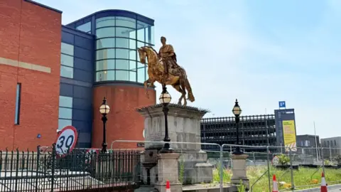 The King Billy statue of the king on a rearing horse in gold with signs of weathering it stands on a stone plinth and is surrounded by steel fencing and road warning signs